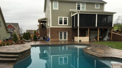 Pool with stone coping and patio, beside a light-gray house. Photo