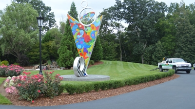A colorful, abstract sculpture sits in a landscaped area, next to a road with a light-colored vehicle. Photo