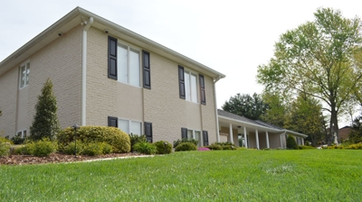 A light beige house sits on a lawn with green trees. Photo
