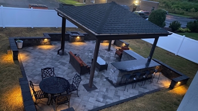stone patio that is covered in the middle of the backyard with white fence, furniture, stone walls and stone counter. Photo