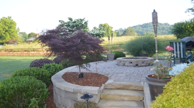 Patio area with small stone fire pit, furniture, tree in mulch and stone steps leading down to the grass and bushes in mulch. Photo