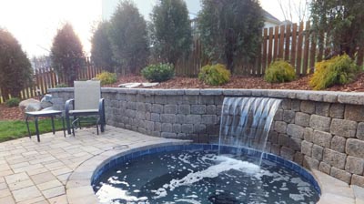 Round hot tub with stone walls, waterfall feature and patio furniture. Photo